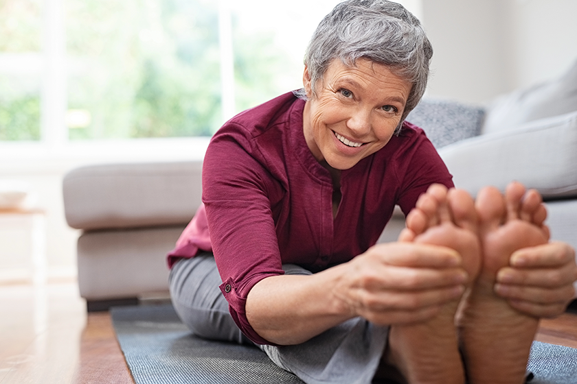 Closeup of senior woman stretching to touch toes while sitting on yoga mat. Portrait of mature woman doing her stretches at home while looking at camera. Happy healhty lady doing yoga exercises. 