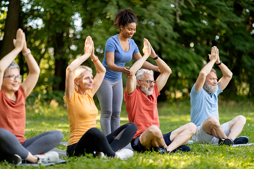 Group of active senior people in sportrswear attending outdoor yoga class with instructor, mature friends meditating with hands above head, sitting on fitness mats, enjoying healthy lifestyle